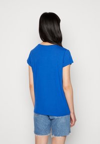 Modrá košile s krátkým rukávem vyrobená z měkkého materiálu, s kulatým výstřihem. Záda jsou jednoduchá, bez jakýchkoli vzorů nebo kovových detailů.