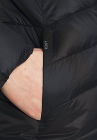 Svart vadderad jacka med quiltat mönster och en sidoficka med en svart tygetikett märkt "ROXY." Slät, glänsande yta.