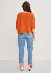 Suéter naranja de punto con mangas anchas, combinado con jeans de color azul claro, enrollados y zapatillas multicolor, visto desde la espalda.