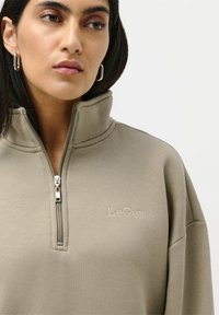 Kvinne med mørkt hår har på seg sølvfargede hoopsmykker og en beige zip-up hettegenser med "LeGer" brodert på brystet.