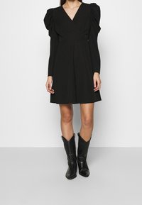 Schwarzes Wickelkleid mit langen Puffärmeln, V-Ausschnitt und ausgestelltem Rock. Kombiniert mit kniehohen schwarzen Cowboy-Stiefeln mit dekorativer Naht.