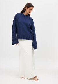 Navy knit sweater with ribbed cuffs and hem, paired with a white ribbed maxi skirt and strappy sandals, set against a neutral background.