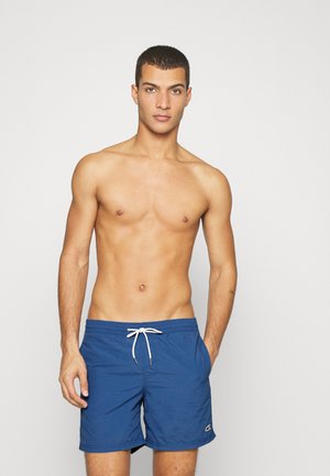 Blaue Badehose mit Kordelzugbund, Seitentaschen und kleinem Logo am Saum. Glatte Textur, Design bis zur Mitte des Oberschenkels.