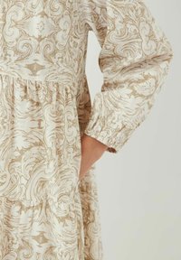 Langarm jurk in crème met een draaiend beige patroon. Heeft een geplooide taille en elastische manchetten. Inclusief zijzakken.
