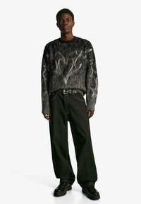 Fekete és szürke, oversized kötött pulóver, változatos textúrákkal, világosszürke örvénymintával. Széles lábú fekete farmernadrággal és fekete cipőkkel párosítva.