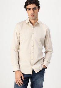 Cămașă bej cu nasturi, guler și mâneci lungi, având o textură netedă și o ușoară strălucire, asortată cu blugi din denim închis.