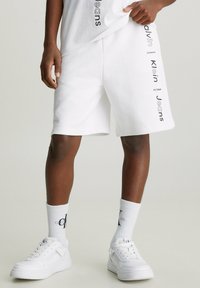 Witte katoenen shorts met zwart bedrukte "Calvin Klein Jeans" tekst aan de zijkanten, gecombineerd met witte sneakers en logo sokken. Soepele textuur.