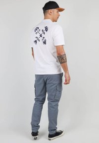 T-shirt blanc avec un motif floral bleu marine dans le dos, associé à un pantalon cargo gris et des baskets noires avec des accents blancs.