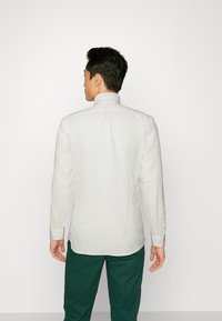 Camisa blanca de manga larga con cuello clásico, tejido texturizado y puños abotonados, combinada con pantalones verdes. Vista desde la parte posterior.