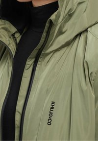 Olivgrön lättviktsjacka med huva, med en svart dragkedja och logotypen "KHUJO CO" i en kontrasterande svart font på sidan.