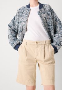 Bomber jacket con motivo floreale in toni di blu, con polsini navy, abbinato a pantaloni corti beige plissettati fino al ginocchio. Sono visibili morbide trame di cotone.