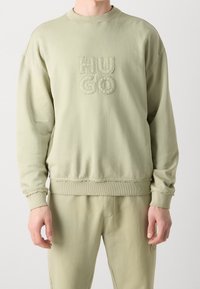 Man die een lichtgroene trui en bijpassende broek draagt met "HUGO" geborduurd op de borst, staand tegen een witte achtergrond.