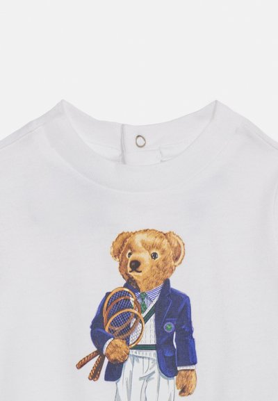 Chemise blanche avec un ours en peluche habillé d'un blazer bleu, d'une chemise rayée, d'une cravate verte et d'un pantalon blanc, tenant deux raquettes de tennis marron.
