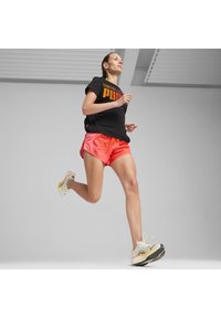 Camiseta atlética negra con texto naranja, pantalones cortos rosas con un patrón y zapatillas de correr de color claro con una suela texturizada, en movimiento.