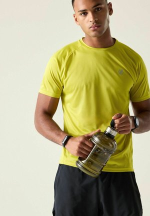 Joven con camiseta deportiva amarilla sosteniendo una botella de agua grande, lleva pulseras de actividad en ambas muñecas, de pie frente a un fondo claro y sencillo.