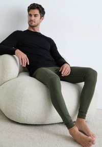 Black long-sleeve shirt and olive green leggings, fitted design, smooth texture, seated on a white, soft-textured chair against a neutral backdrop.