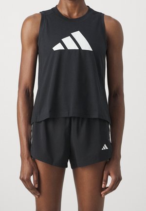 Osoba nosící černý sportovní nátělník bez rukávů a k tomu odpovídající krátké kalhoty, obojí s bílým logem Adidas na přední straně.