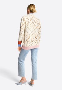 Gestructureerde crème cardigan met bruine spikkels, een roze geribde zoom en meerkleurige mouwen. Gecombineerd met lichtblauwe jeans en grijze hakken.