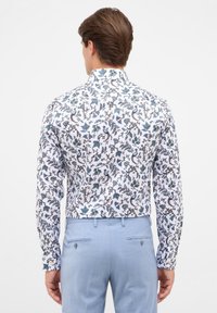 Witte shirt met intricaties in blauw, groen en bruine bloemenpatronen; lange mouwen, klassieke kraag, gladde katoenen stof; gedragen met lichtblauwe broek.