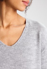 Pull en maille grise avec un col en V profond, une texture douce et une coupe décontractée, mettant en valeur une apparence épurée et minimaliste.
