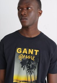 Camiseta negra con un gráfico tropical impreso. Presenta el texto amarillo "GANT Resort" y palmeras contra un fondo de atardecer. Material de algodón suave.