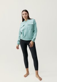 Jasnoniebieska kurtka zapinana na zamek błyskawiczny z dwiema kieszeniami na piersi i elastycznymi mankietami, w połączeniu z czarnymi legginsami. Gładka tekstura i dopasowany krój.