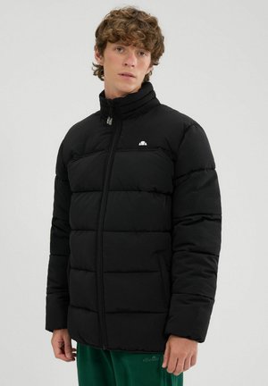 Joven con una chaqueta acolchada negra de cuello alto y pantalones verdes, de pie frente a un fondo claro y liso.