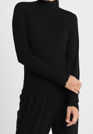 Mujer con una blusa ajustada de manga larga negra con bordes en forma de concha en los puños y cuello alto, combinada con pantalones negros sueltos.