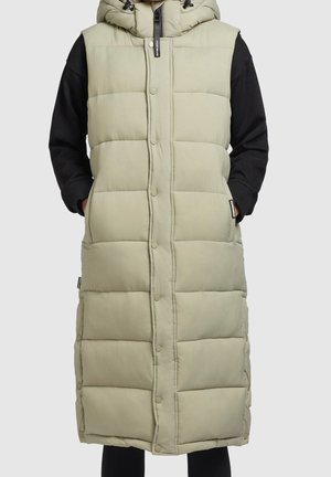 Długa beżowa pikowana kamizelka bez rękawów z zatrzaskami, kapturem i kieszeniami po bokach, noszona na czarnym topie z długim rękawem.