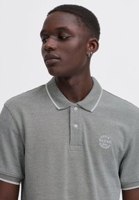 Junger Mann mit einem hellgrauen Poloshirt mit weißem Abschluss und kleinem Brustlogo, blickt mit neutralem Gesichtsausdruck nach links.