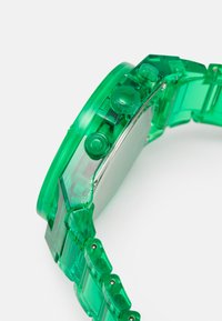Przezroczysty zielony plastikowy zegarek z okrągłą tarczą, okrągłymi guzikami i segmentowym paskiem. Posiada stalowy tył i gładką teksturę.
