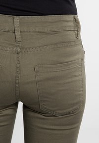 Olive zaļas džinses ar elegantu piegriezumu, kurām ir divas aizmugures kabatas, smalks diegs un gluda tekstūra.