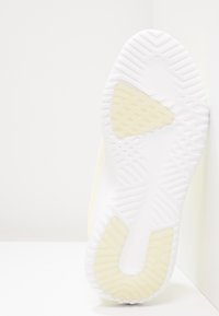 Vit sportskosula med texturerat mönster och gula accenter. Böjd form med mönstrad yta och utskärningar.