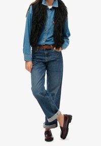 Koszula z denimu z kołnierzykiem, czarna kamizelka z futerka ekologicznego, niebieskie jeansy z wysokim stanem, brązowy pleciony pasek i ciemnoczerwone mokasyny na wsuwkę.