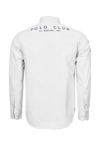 Bílá košile s dlouhým rukávem a manžetami na knoflíky, s tmavě modrým vyšívaným textem „POLO CLUB LA MARTINA 1987“ na zádech. Hladká textura.