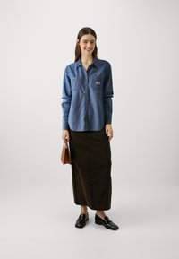 Denim button-up shirt with two chest pockets, paired with a brown long skirt and black loafers, complemented by a small brown handbag.