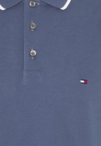Polo bleu texturé avec trois boutons foncés et un petit logo rectangulaire rouge, blanc et marine sur le côté droit.