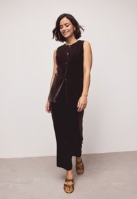 Mustast sametist varrukateta pluus, millel on nööbid, kombineeritud pika musta seelikuga. Komplekt on pehme tekstuuri ja minimalistliku disainiga. Pruunid sandaalid vibu detailidega.