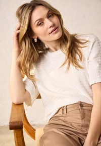 Femme aux longs cheveux blonds portant un haut blanc texturé à manches courtes et un pantalon beige, appuyée sur une chaise en bois avec une expression détendue.