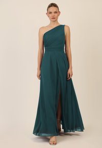 Türkises, einseitig getragenes Abendkleid mit gefaltetem Oberteil und einem Schenkelhohen Schlitz. Hergestellt aus fließendem Stoff mit glatter Textur. Silberne High Heels sind sichtbar.