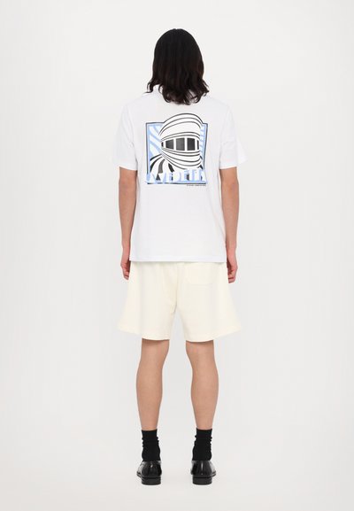 T-shirt blanche avec un graphique dans le dos représentant un design de casque en noir et bleu, porté avec un short clair et des chaussettes.
