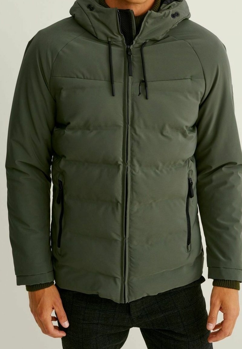 Mężczyzna w oliwkowozielonej pikowanej kurtce z kapturem, zapinanej na czarny zamek błyskawiczny, z zamykanymi bocznymi kieszeniami, noszący ciemne spodnie w kratę.