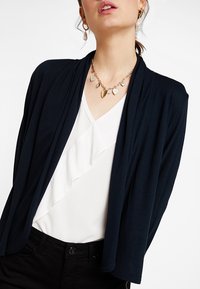 Marinblå omslagskofta i lätt material med öppen framsida. I kombination med en vit blus med volanger och ett guldhänge. Svarta jeans syns.