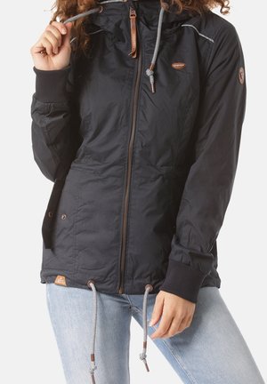 Woman wearing a black zip-up jacket with white drawstrings, ribbed cuffs, side pockets, and light blue jeans against a plain background.