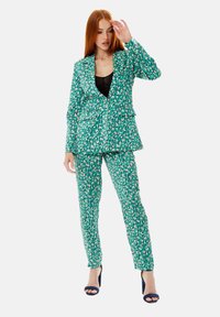 Green patterned suit with a blazer and fitted trousers, featuring abstract shapes in pink and white. Smooth fabric texture, tailored design.
