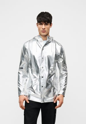 Mand iført skinnende sølvfarvet hættetrøje regnjakke med forlommer og sorte bukser, stående mod en ensartet lys baggrund.