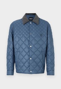 Gesteppte blaue Jacke mit grauem Kragen, Druckknöpfen und einem dezenten Emblem. Mit Rautenmuster und seitlichen Taschen.