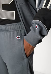 Šedé tepláky s elastickým pásom a zúženými členkami, ktoré majú nášivku s logom na pravej kapse a kontrastné čierne akcenty na vrchu.