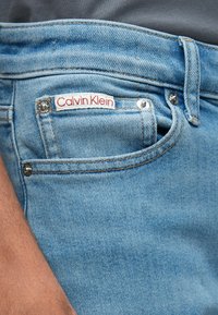 Svetlo modre kavbojke iz jeansa s sprednjim žepom, na pasu pa je pritrjena bela etiketa z rdečim napisom "Calvin Klein".