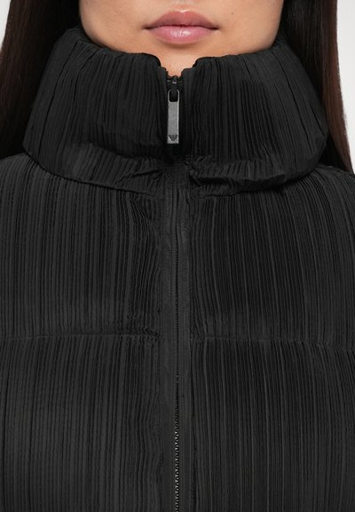 Black pleated jacket with high collar and zip closure. Features textured fabric and ribbed details. Design is form-fitting and modern.
